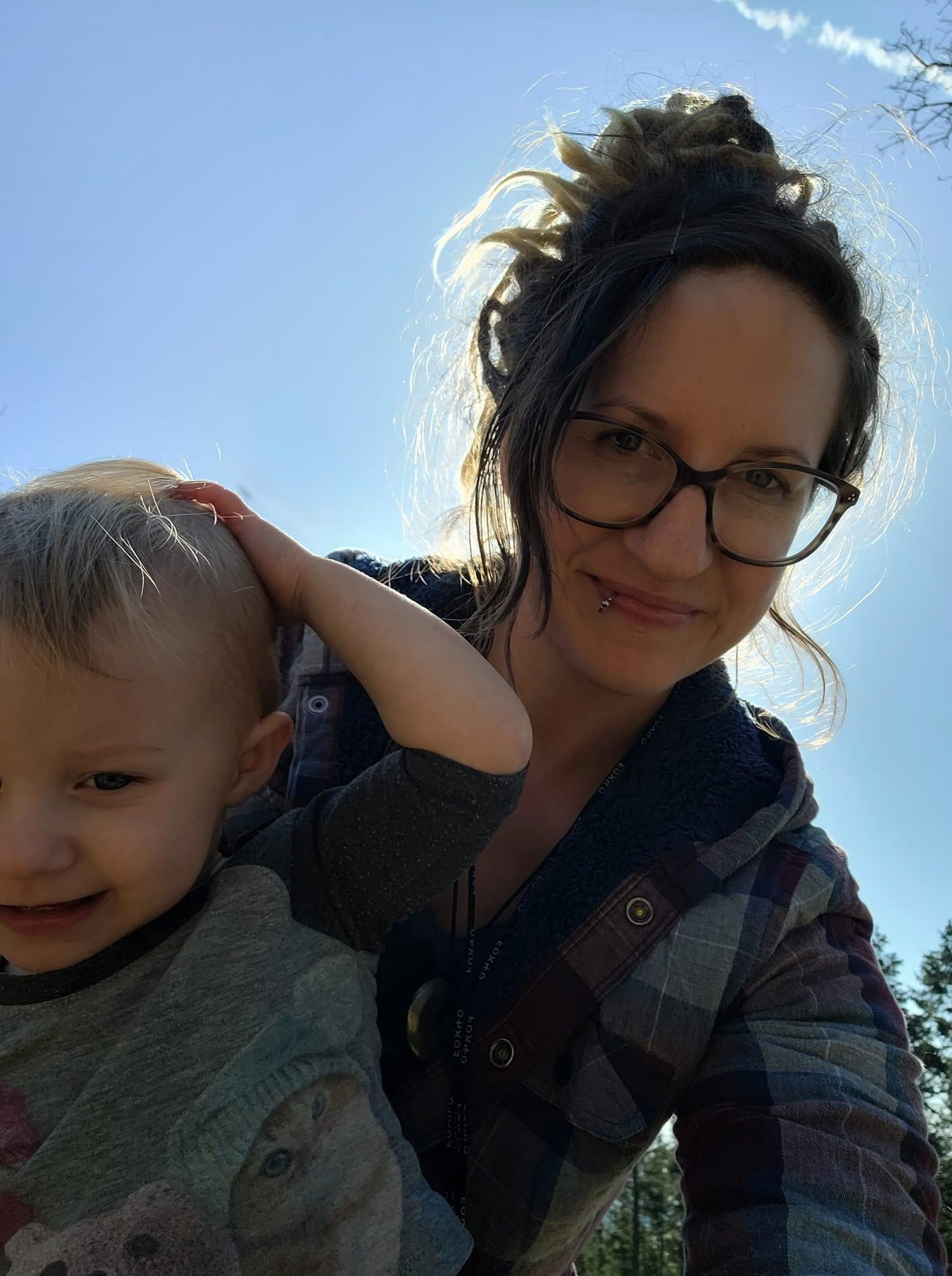 A lovely lady with dark hair and large dark rimmed glasses silhouetted against the sun holding her smiling toddler child